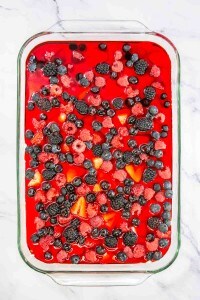 A glass dish brimming with red gelatin is crowned with a medley of berries—raspberries, blackberries, and blueberries—evoking the sweet charm of a Mixed Berry Pretzel Salad, all elegantly displayed on a cool marble surface.