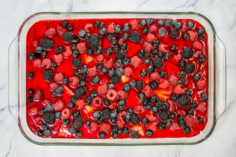 A glass dish brimming with a red gelatin mixed berry pretzel salad is topped with blackberries, blueberries, and raspberries, elegantly set on a marble surface.