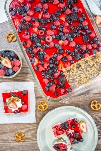 A glass dish with mixed berry pretzel salad, featuring a berry-topped red gelatin dessert, partially served on a wooden table alongside scattered pretzels and a small bowl of mixed berries.