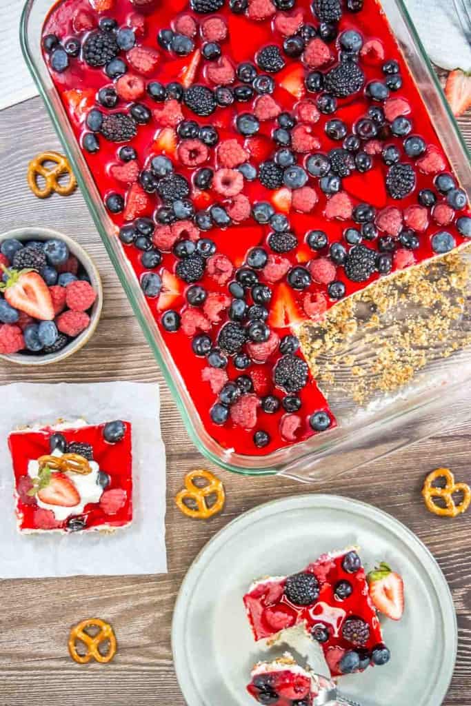 A glass dish with mixed berry pretzel salad, featuring a berry-topped red gelatin dessert, partially served on a wooden table alongside scattered pretzels and a small bowl of mixed berries.