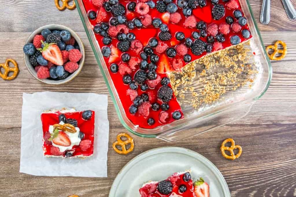 A glass dish showcases a Mixed Berry Pretzel Salad, the red gelatin dessert adorned with berries. A slice is missing, resting on a small plate next to a bowl of berries and scattered pretzels on the wooden table.