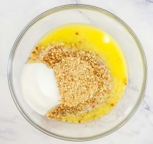 A glass bowl holds a delightful mix of melted butter, chopped nuts, sugar, and crushed graham crackers on a marble surface, reminiscent of the crunchy base one might find in a mixed berry pretzel salad.