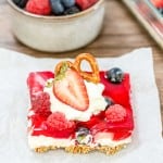 A dessert square inspired by a Mixed Berry Pretzel Salad, featuring a graham cracker crust, creamy layer, and red gelatin topping. It's garnished with whipped cream, a pretzel twist, and fresh berries. A bowl of mixed berries complements from the background.