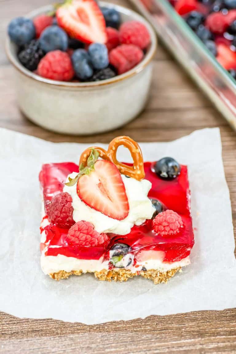A dessert square inspired by a Mixed Berry Pretzel Salad, featuring a graham cracker crust, creamy layer, and red gelatin topping. It's garnished with whipped cream, a pretzel twist, and fresh berries. A bowl of mixed berries complements from the background.