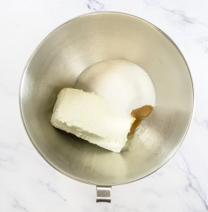 Cream cheese, sugar, and vanilla extract in a stainless steel mixing bowl on a marble countertop, ready to transform into a delightful mixed berry pretzel salad.