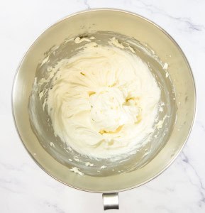 A stainless steel bowl containing whipped cream for a delightful Mixed Berry Pretzel Salad sits on a white marble surface.