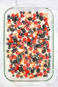 A glass dish featuring a Mixed Berry Pretzel Salad, topped with strawberries, blueberries, raspberries, and blackberries on a light cream base.