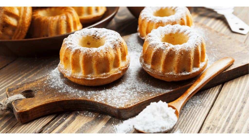 Mini Bundt cakes dusted with powdered sugar on a wooden cutting board, with a wooden spoon of sugar nearby.