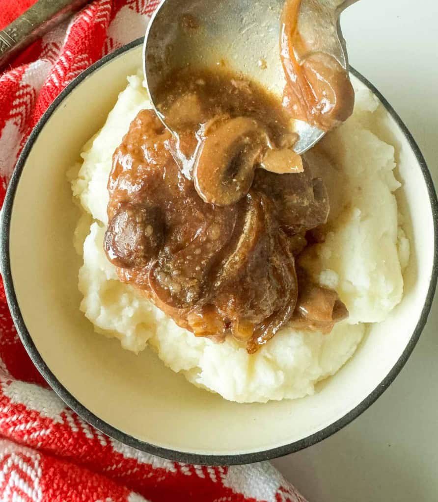 A bowl of mashed potatoes topped with rich mushroom gravy, reminiscent of a slow cooker Salisbury steak, with a spoon serving more gravy. A red and white cloth is partially visible beside the bowl.