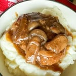 A bowl of mashed potatoes topped with a savory mushroom gravy reminiscent of slow cooker Salisbury steak. A red and white checkered cloth is partially visible next to the bowl, adding a touch of rustic charm.