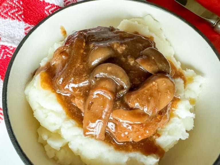 A bowl of mashed potatoes topped with a savory mushroom gravy reminiscent of slow cooker Salisbury steak. A red and white checkered cloth is partially visible next to the bowl, adding a touch of rustic charm.