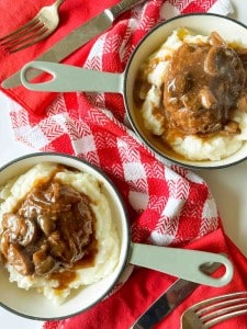 Two bowls of mashed potatoes topped with slow cooker Salisbury steak gravy and mushrooms sit on a red-and-white checkered cloth, forks at the ready.