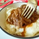 A bowl of mashed potatoes topped with brown gravy is being mixed with a fork, reminiscent of a slow cooker Salisbury steak meal. A red and white cloth and utensils are set in the background.