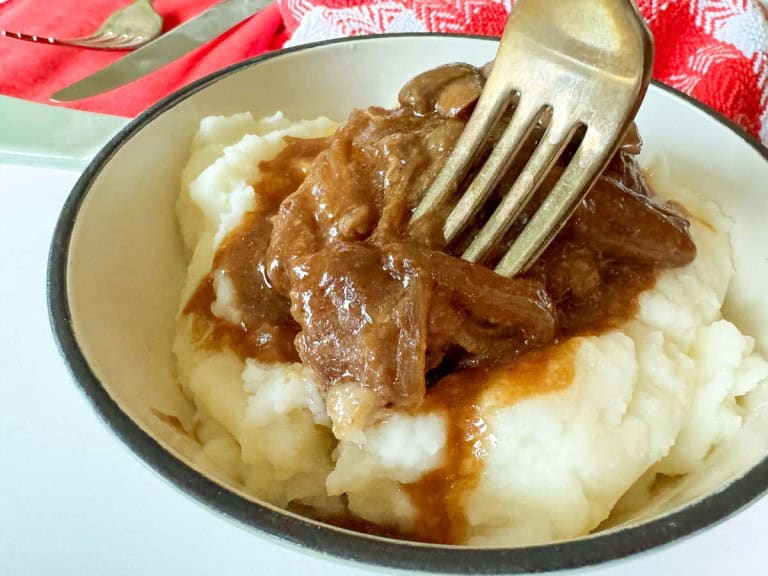 A bowl of mashed potatoes topped with brown gravy is being mixed with a fork, reminiscent of a slow cooker Salisbury steak meal. A red and white cloth and utensils are set in the background.