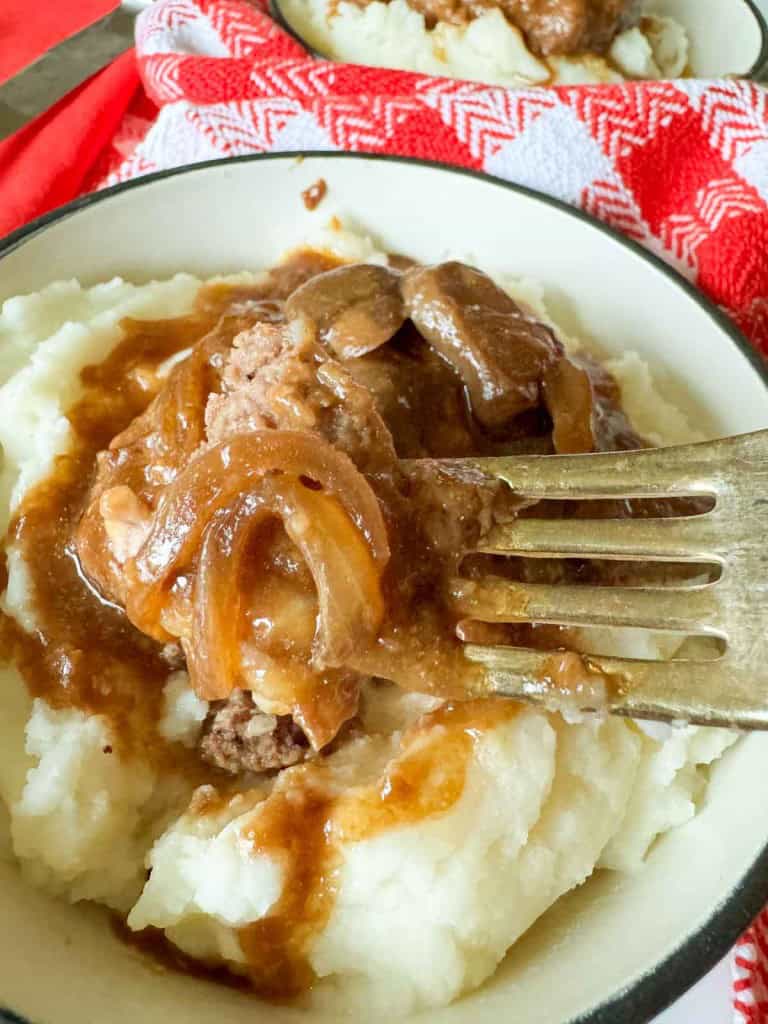 Mashed potatoes topped with a rich meat and onion gravy reminiscent of slow cooker Salisbury steak, with a fork resting on the side. A red and white cloth serves as the charming backdrop.