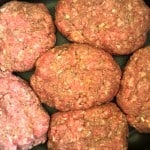 Six raw hamburger patties, seasoned with visible bits of onions, are arranged on a dark surface ready for cooking&mdash;perfect for transforming into succulent slow cooker Salisbury steak.