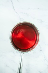 A stainless steel saucepan filled with red liquid, perfect for making strawberry pretzel salad, sits on a white marble surface.