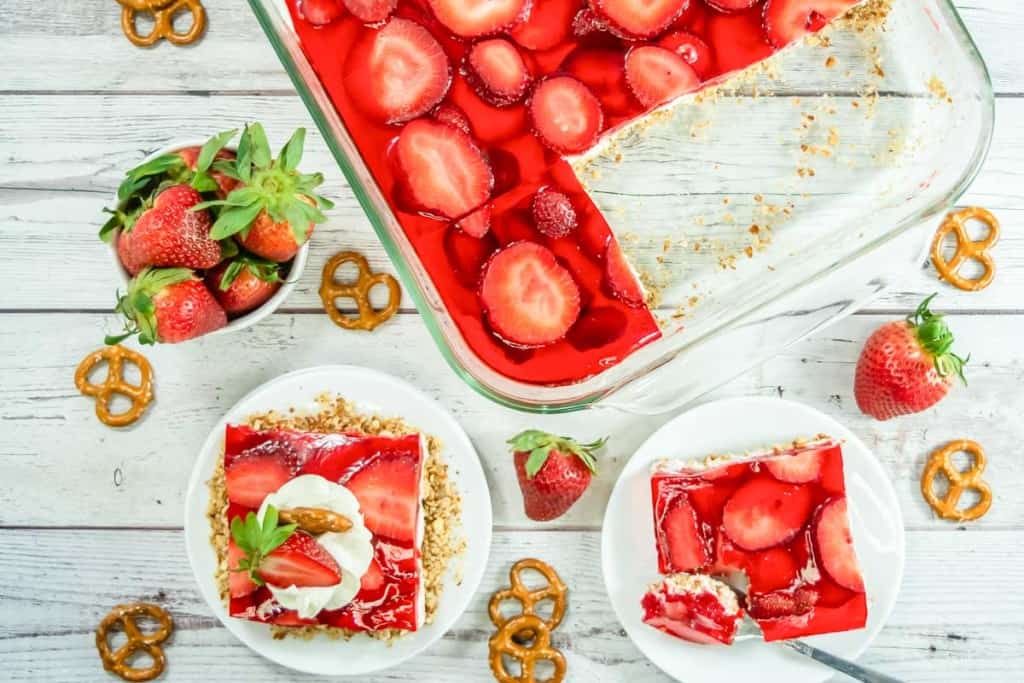 Overhead view of strawberry pretzel salad in a glass dish, with two plated servings, fresh strawberries, and pretzels arranged on a white wooden surface.