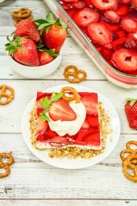 A slice of strawberry pretzel salad dessert topped with whipped cream, a half strawberry, and a pretzel on a white plate, with a bowl of strawberries and a tray of strawberry pretzel salad in the background.