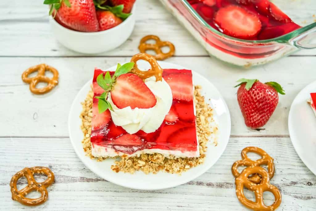 A slice of strawberry pretzel salad topped with whipped cream, a strawberry, and a pretzel on a white plate, with extra pretzels and strawberries scattered around.