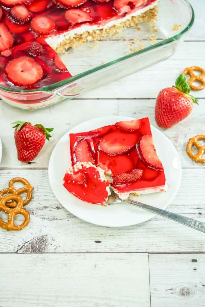 A slice of strawberry pretzel salad sits on a white plate with a fork, surrounded by fresh strawberries and pretzels, while a dish of the irresistible strawberry pretzel salad rests in the background.