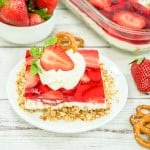 A slice of strawberry pretzel salad dessert topped with whipped cream and a fresh strawberry, served on a white plate with pretzels and whole strawberries nearby.