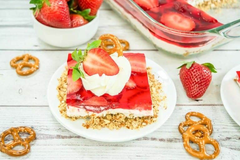 A slice of strawberry pretzel salad dessert topped with whipped cream and a fresh strawberry, served on a white plate with pretzels and whole strawberries nearby.