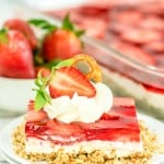 A slice of strawberry pretzel salad dessert topped with whipped cream, a pretzel, and a strawberry half, served on a white plate with more strawberry pretzel salad and fresh strawberries in the background.