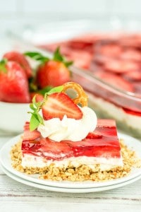 A slice of strawberry pretzel salad dessert topped with whipped cream, a pretzel, and a strawberry half, served on a white plate with more strawberry pretzel salad and fresh strawberries in the background.