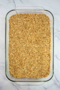 A glass baking dish filled with an even layer of oat and crumb mixture, reminiscent of the crust in a classic strawberry pretzel salad, sits on a white marble surface.