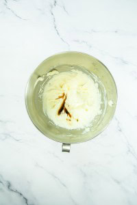 A stainless steel mixing bowl containing whipped cream cheese and vanilla extract sits on a white marble surface, ready to become the creamy layer for a classic strawberry pretzel salad.