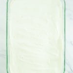 A glass baking dish filled with strawberry pretzel salad, topped with a smooth layer of white frosting, sits on a white marble surface.