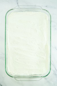 A glass baking dish filled with strawberry pretzel salad, topped with a smooth layer of white frosting, sits on a white marble surface.