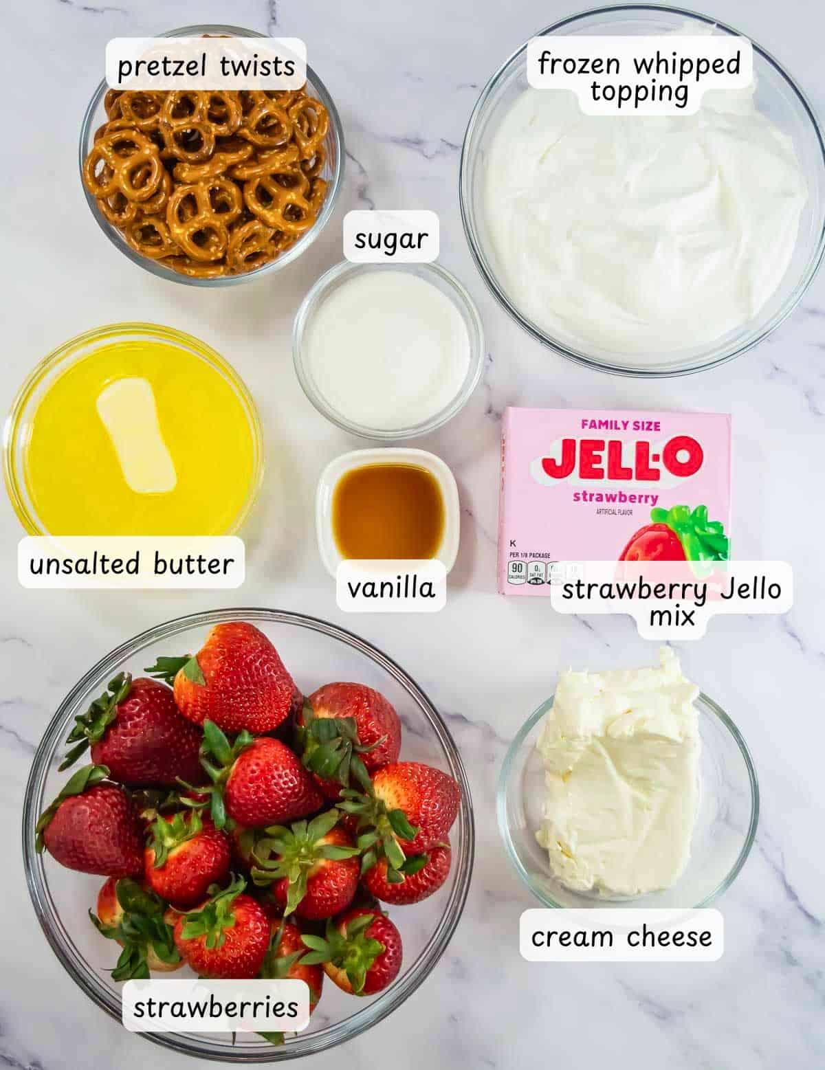 Ingredients for a classic strawberry pretzel salad are arranged on a countertop, including pretzel twists, whipped topping, sugar, butter, vanilla, strawberry Jello mix, strawberries, and cream cheese.