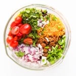 A glass bowl containing taco pasta salad ingredients like chopped red onion, grape tomatoes, shredded cheese, cooked ground meat, diced green pepper, and fresh cilantro, viewed from above.