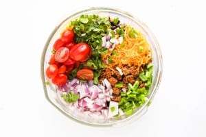 A glass bowl containing taco pasta salad ingredients like chopped red onion, grape tomatoes, shredded cheese, cooked ground meat, diced green pepper, and fresh cilantro, viewed from above.