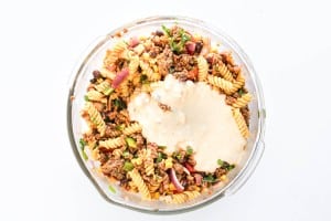 A glass bowl filled with taco pasta salad—rotini pasta, ground beef, beans, red onions, and herbs—all topped with a creamy white sauce.