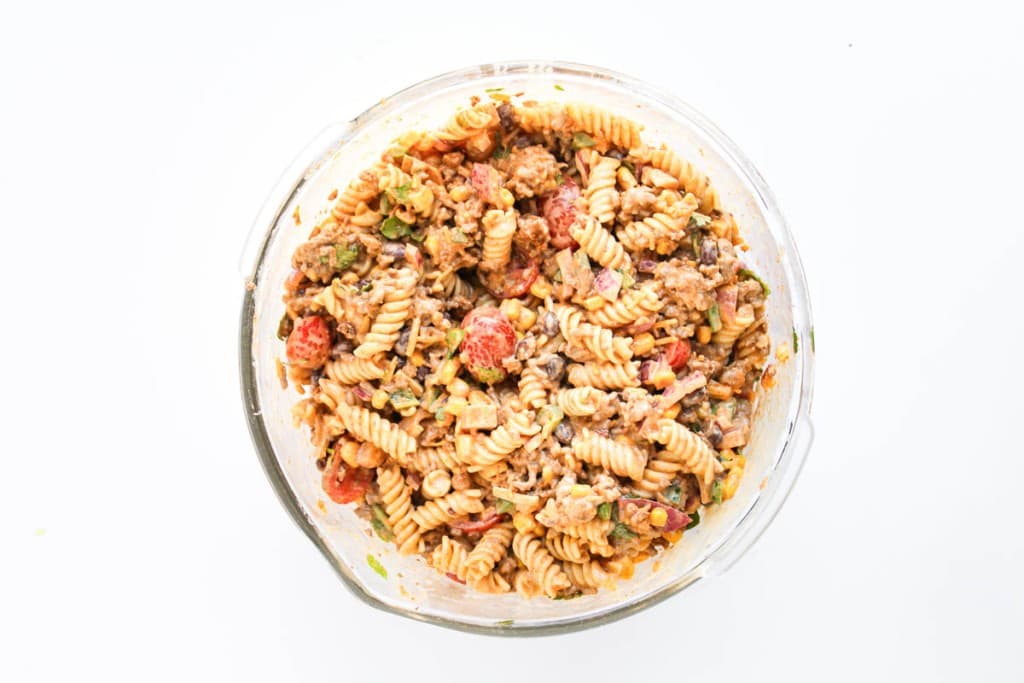 A glass bowl filled with taco pasta salad—rotini pasta mixed with ground meat, black beans, corn, tomatoes, and chopped green herbs.