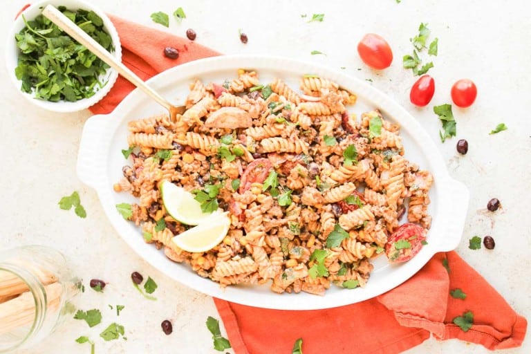 A white oval dish filled with taco pasta salad featuring rotini, ground meat, cherry tomatoes, and herbs, garnished with lime wedges and chopped cilantro, sits on an orange napkin.