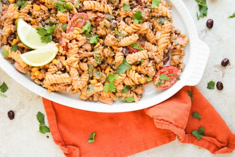 A white dish filled with taco pasta salad—rotini, ground meat, corn, black beans, tomatoes, and cilantro—garnished with lime wedges sits on an orange napkin.