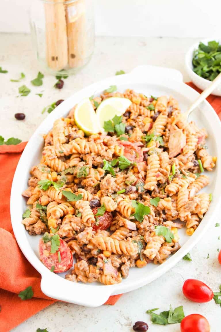 A white oval dish filled with taco pasta salad made of rotini pasta, ground meat, black beans, tomatoes, and herbs, all garnished with cilantro and lemon wedges.