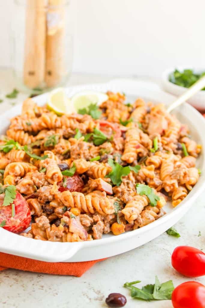 A white dish filled with taco pasta salad featuring rotini, ground meat, tomato slices, creamy sauce, and fresh cilantro garnish, served with lime wedges on the side.