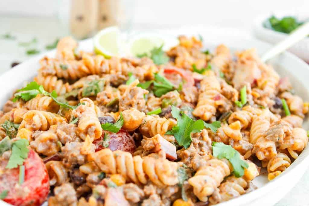 A plate of taco pasta salad featuring rotini mixed with ground meat, tomatoes, corn, and garnished with fresh cilantro.