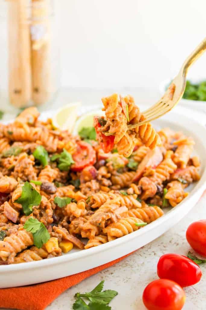 A white dish filled with creamy taco pasta salad, rotini, vegetables, and herbs; a fork is lifting a bite above the plate. Cherry tomatoes and cilantro are nearby.