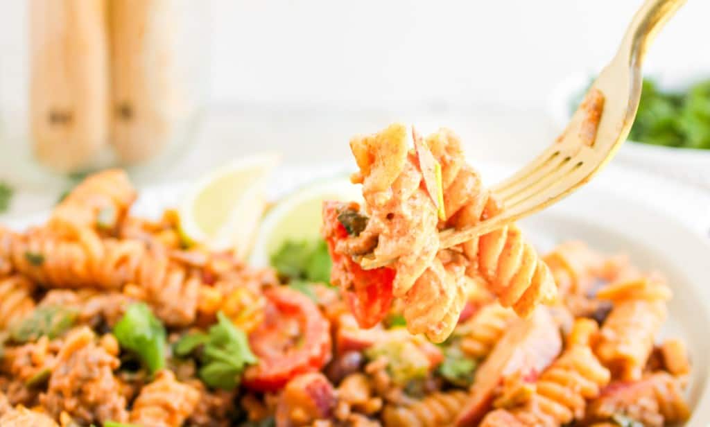 A fork holds a bite of taco pasta salad with rotini, sauce, vegetables, and herbs above a plate of the same flavorful dish.