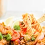 A fork holds a bite of taco pasta salad with rotini, sauce, vegetables, and herbs above a plate of the same flavorful dish.