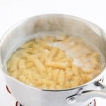 A stainless steel pot filled with boiling water and rotini pasta, perfect for making taco pasta salad, sits on a patterned trivet against a white background.
