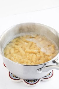 A stainless steel pot filled with boiling water and rotini pasta, perfect for making taco pasta salad, sits on a patterned trivet against a white background.