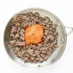 A stainless steel pan with browned ground beef and a pile of taco seasoning powder in the center, viewed from above on a white background—perfect for starting a flavorful taco pasta salad.