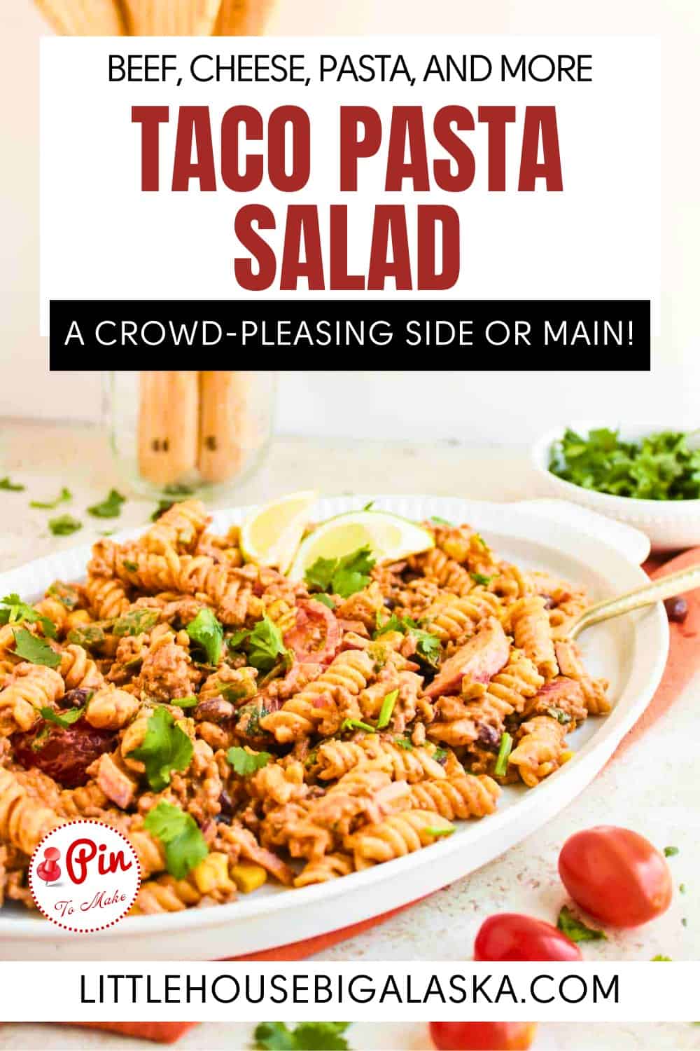 A serving dish filled with taco pasta salad, garnished with herbs, is displayed on a table with serving utensils and fresh ingredients nearby.